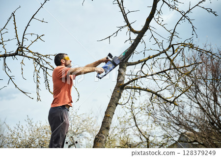 Young man cutting trees using electrical chainsaw in motion at his backyard 128379249
