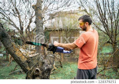 Young man cutting trees using electrical chainsaw in motion at his backyard Young man cutting trees using electrical chainsaw in motion at his backyard 128379251