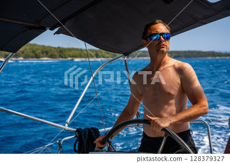 Man with bare torso steering a yacht on the open sea on a sunny day. Concept: summer vacation, yachting, sea travel. Man with bare torso steering a yacht on the open sea on a sunny day. Concept: summer vacation, yachting, sea travel. 128379327