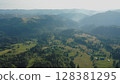 Aerial view of a fog creeping over the mountains surrounding the valley on a summer day in the morning. Aerial drone view of houses in a mountain valley on a summer day. 128381295