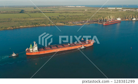 Aerial view of a large cargo ship leaving the harbor with tugboats at sunset. On the horizon in the roadstead there are ships waiting for the entrance to the port. 128381299