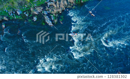 Aerial view of a kayak boat rushing along with a stormy stream between the stones at high speed. Rowers trying to steer the boat in the right direction. 128381302
