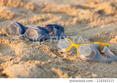 Pair of swim goggles lying on sandy beach with warm evening light 128383515