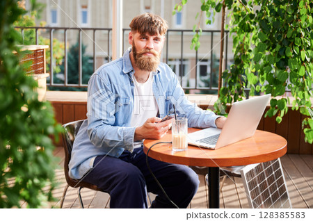 Man works on laptop and smartphone at outdoor table, with glass of iced coffee. Solar panel nearby, emphasizing sustainable, modern eco-friendly workspace that combines technology and sustainability. Man works on laptop and smartphone at outdoor table, with glass of iced coffee. Solar panel nearby, emphasizing sustainable, modern eco-friendly workspace that combines technology and sustainability. 128385583