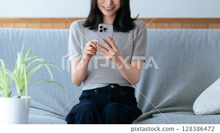 Smiling woman sitting on sofa and using smartphone 128386472