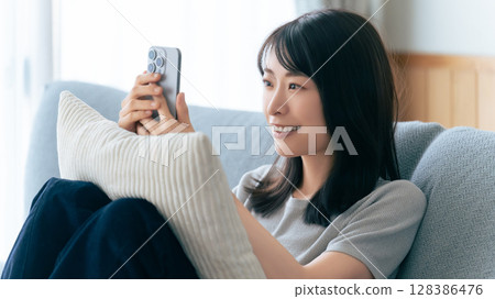 Smiling woman sitting on sofa and using smartphone 128386476