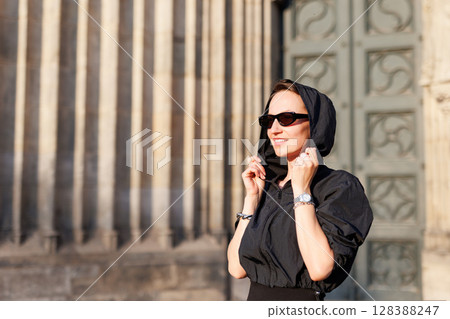 Portrait young adult elegant woman black dress and sunglasses smiles confidently in golden sunlight warm sunset backlit light against historic cathedral building. Female person walking city street Portrait young adult elegant woman black dress and sunglasses smiles confidently in golden sunlight warm sunset backlit light against historic cathedral building. Female person walking city street 128388247