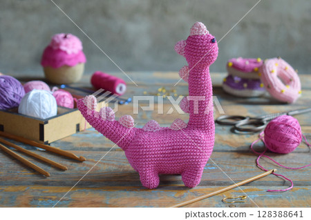 Close up of finished pink amigurumi dinosaur toy on rustic painted wooden table with wool yarn accessories and colorful thread balls crochet craft concept 128388641