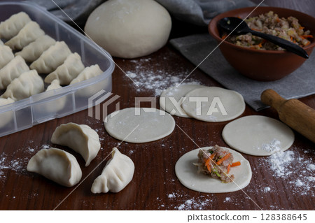 Stuffing pork gyoza with vegetable filling on raw dough rounds folding dumplings on floured wooden board home cooking traditional Japanese meal step by step process in kitchen close up Stuffing pork gyoza with vegetable filling on raw dough rounds folding dumplings on floured wooden board home cooking traditional Japanese meal step by step process in kitchen close up 128388645