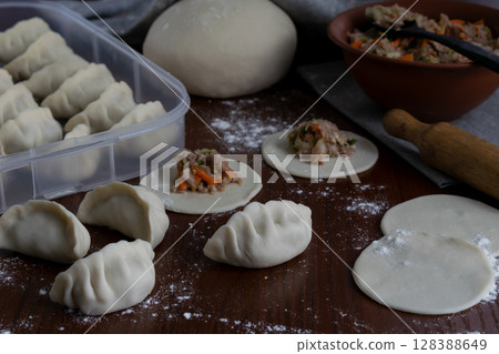 Several raw pork gyoza dumplings being folded on floured wooden table dough circles and filling in progress visible during Japanese cooking at home traditional meal preparation view 128388649