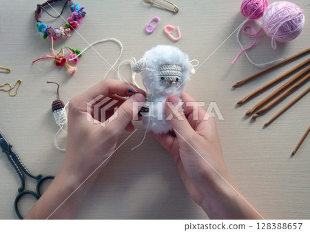 Handmade soft toy process with hands knitting small plush animal using crochet hook wool yarn, creative hobby craft table scene close up step by step DIY project, Step 2 - to sew all details of toy 128388657
