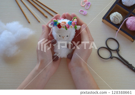 Crocheted sheep toy with floral crown held by female hands after completion DIY amigurumi soft animal project on desk with yarn and scissors visible finished handmade figure 128388661
