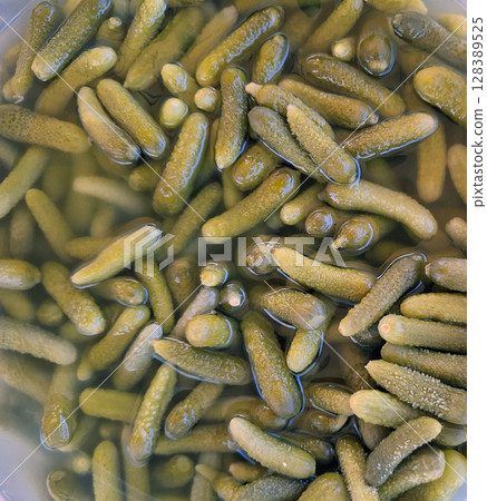 Small pickled cucumbers, also known as gherkins, are densely packed and submerged in clear brine, highlighting their glossy texture and green color in a close-up view. 128389525