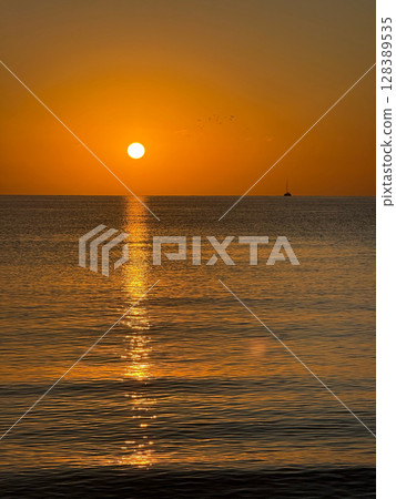 A breathtaking sunset over the calm sea with a sailboat on the horizon. Warm orange and golden tones fill the sky and reflect on the water. Travel, nature, relaxation, and maritime concept A breathtaking sunset over the calm sea with a sailboat on the horizon. Warm orange and golden tones fill the sky and reflect on the water. Travel, nature, relaxation, and maritime concept 128389535