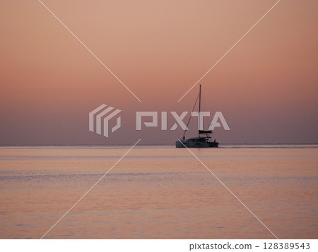 A breathtaking sunset over the calm sea with a sailboat on the horizon. Warm orange and golden tones fill the sky and reflect on the water. Travel, nature, relaxation, and maritime concept 128389543