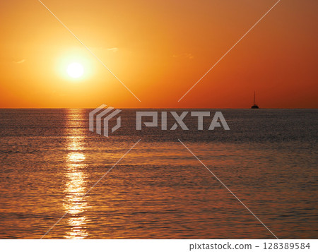 A breathtaking sunset over the calm sea with a sailboat on the horizon. Warm orange and golden tones fill the sky and reflect on the water. Travel, nature, relaxation, and maritime concept 128389584