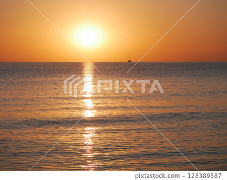A breathtaking sunset over the calm sea with a sailboat on the horizon. Warm orange and golden tones fill the sky and reflect on the water. Travel, nature, relaxation, and maritime concept 128389587