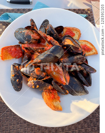 Delicious serving of mussels in spicy tomato sauce complemented by crispy toasted bread slices, served on a white plate, perfect for seafood enthusiasts and culinary photography. 128391103