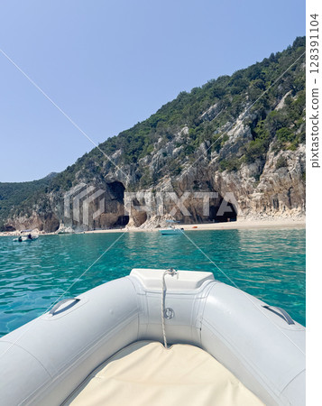 Inflatable boat on vibrant turquoise water approaching rocky sea caves. The pristine shoreline and lush cliffs in the distance create a stunning backdrop for a serene adventure. Cala Luna Sardinian  128391104