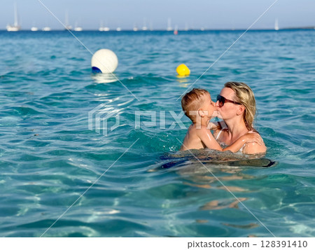 A heartwarming scene of a mother hugging and kissing her young child in a serene ocean setting. Both smile joyfully, embodying love and connection amid gentle waves A heartwarming scene of a mother hugging and kissing her young child in a serene ocean setting. Both smile joyfully, embodying love and connection amid gentle waves 128391410