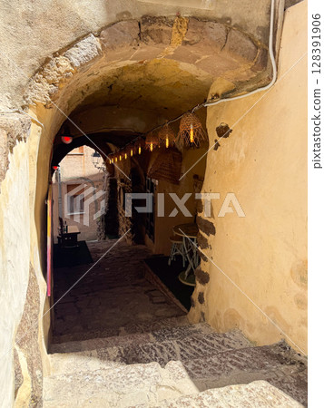Narrow stone pathway beneath an ancient archway adorned with glowing rustic lanterns. The texture of the weathered walls adds a historic ambiance, inviting exploration. 128391906