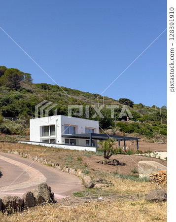 Modern white house nestled in scenic countryside with a winding path leading up a gentle hill. Surrounded by lush greenery, the structure exudes contemporary architectural style. 128391910