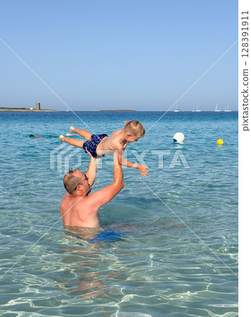Joyful scene of a father lifting his son in the clear sea water. Both are smiling, enjoying a playful moment under the bright sun. Boats and buoys are visible in the background. 128391911