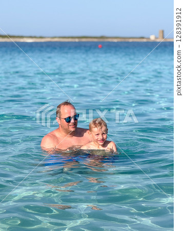 Father and son bond while swimming in clear blue waters. Both are smiling and enjoying the refreshing ocean. The backdrop features a beautiful coastline under a clear sky. 128391912