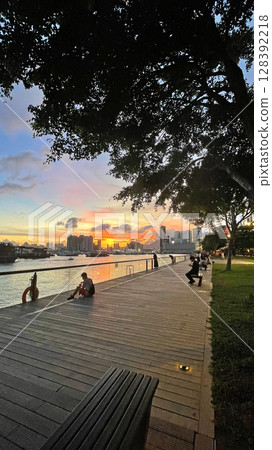 Silhouette of people, park, building, boats on bay water at sunset 128392218