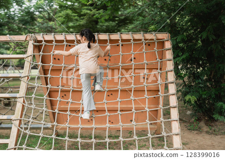 Child Climbing a Vertical Playground Net in a Park Surrounded by Lush Green Trees on a Sunny Day, Joyful Outdoor Activity for Kids 128399016