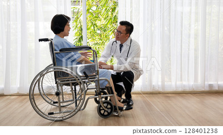 Doctor kneeling beside elderly female patient in wheelchair offering support in hospital room with natural daylight through white curtains Doctor kneeling beside elderly female patient in wheelchair offering support in hospital room with natural daylight through white curtains 128401238