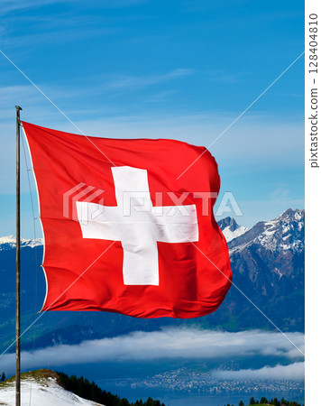 Switzerland flag waving against mountain backdrop in bright blue sky with clouds during daytime Switzerland flag waving against mountain backdrop in bright blue sky with clouds during daytime 128404810