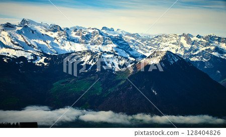 Majestic snow-capped mountains rise above serene green valleys under a clear blue sky in the Swiss Alps during midday 128404826
