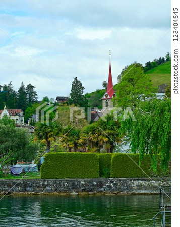 Scenic lakeside view featuring lush greenery and a charming church with a red spire 128404845