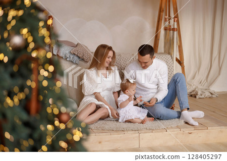 Happy family with baby sitting on cozy rug near Christmas tree in bright wooden room Happy family with baby sitting on cozy rug near Christmas tree in bright wooden room 128405297