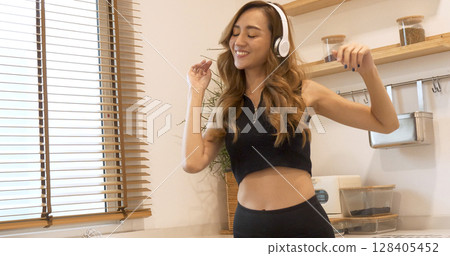 Happy asian dancing woman use headphones listen to music from smartphone sitting on cozy sofa in living room at home. Music mental therapist relaxation woman. Cheerful wellness women dance exercise 128405452