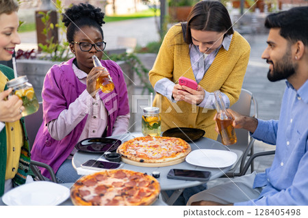 Friends enjoying pizza and drinks at an outdoor cafe, capturing moments with a phone and having a good time together. Friends enjoying pizza and drinks at an outdoor cafe, capturing moments with a phone and having a good time together. 128405498