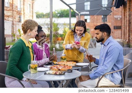 Diverse group of friends enjoying pizza and drinks together at an outdoor restaurant, smiling and using a phone. 128405499