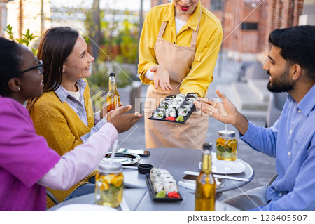 A server presents sushi to a group of friends enjoying a meal outdoors. 128405507