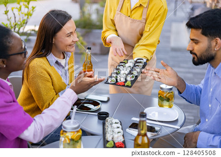 Friends enjoying sushi and drinks at an outdoor restaurant, served by a waiter. Friends enjoying sushi and drinks at an outdoor restaurant, served by a waiter. 128405508