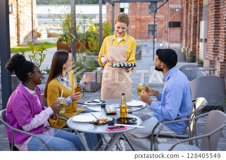 A friendly waitress serves sushi to a group of friends enjoying a meal at an outdoor restaurant. A friendly waitress serves sushi to a group of friends enjoying a meal at an outdoor restaurant. 128405549