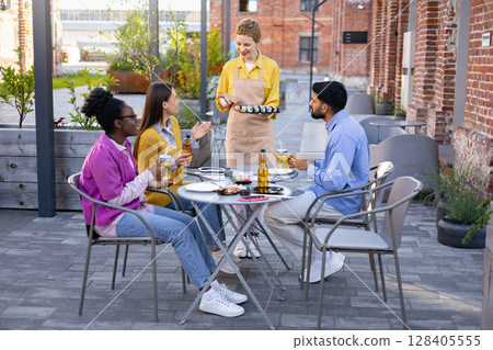 Friends enjoy sushi and drinks outdoors as a server delivers a plate of sushi at a restaurant. Friends enjoy sushi and drinks outdoors as a server delivers a plate of sushi at a restaurant. 128405555