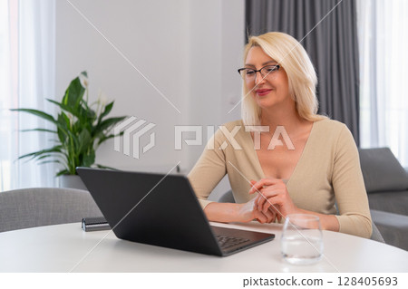 Blonde female psychologist in beige outfit holding online session on laptop. Smiling middle aged therapist conducting remote consultation using notebook in cozy home interior. Mental health and online 128405693