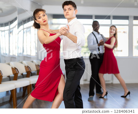 Happy smiling woman dressed in red dress enjoying waltz with male partner during group party in latin dance salon. Social dancing concept Happy smiling woman dressed in red dress enjoying waltz with male partner during group party in latin dance salon. Social dancing concept 128406341