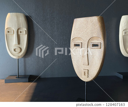 Wooden mask on stand in room. Design in interior. Simple art object in room on dark background. Wooden mask on stand in room. Design in interior. Simple art object in room on dark background. 128407163