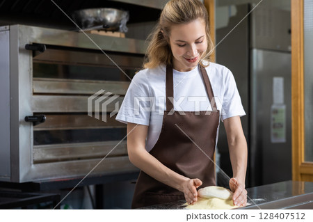 A young woman in a brown apron is preparing dough on a work surface A young woman in a brown apron is preparing dough on a work surface 128407512