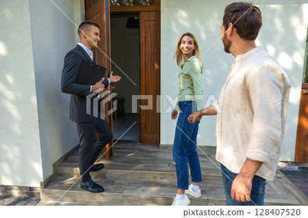 Real estate agent welcoming couple into modern house entrance 128407520