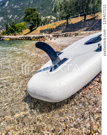 Stand-up paddleboard on pebble beach beside turquoise Lake Garda waters. High quality photo 128407555