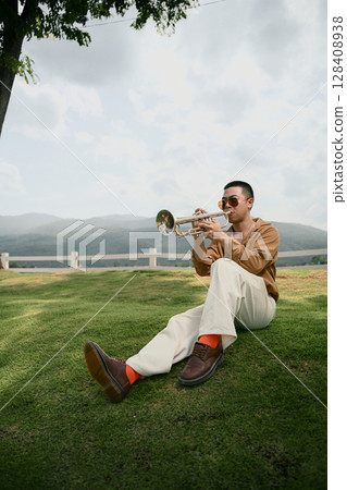 Man sitting on a green lawn playing trumpet with mountain scenery in the background Man sitting on a green lawn playing trumpet with mountain scenery in the background 128408938