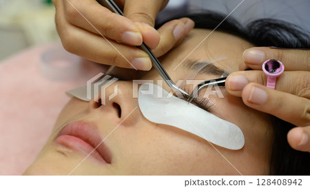 Close up of beauty salon technician applying individual eyelash extensions with precision tweezers Close up of beauty salon technician applying individual eyelash extensions with precision tweezers 128408942